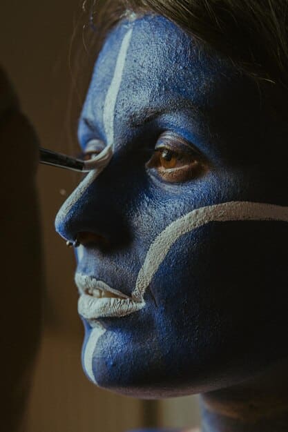 A detailed close-up of a special effects artist applying intricate makeup to an actor. The makeup is designed to create a monster or creature effect, with visible textures, colors, and prosthetics. The artist is focused and precise, highlighting the skill and artistry involved in the process.