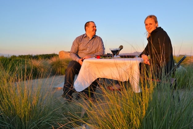 A screenshot of the bachelor on a one-on-one date, laughing and sharing a meal with one of the contestants at a scenic location, such as a vineyard or a beach at sunset.