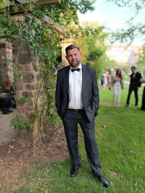 A medium shot showing Connor Roy looking distraught during his wedding reception. In the background, Willa Ferreyra appears concerned, and the opulent wedding decorations are slightly blurred, emphasizing Connor's emotional state. The lighting highlights his anxiety and the underlying tension of the episode.