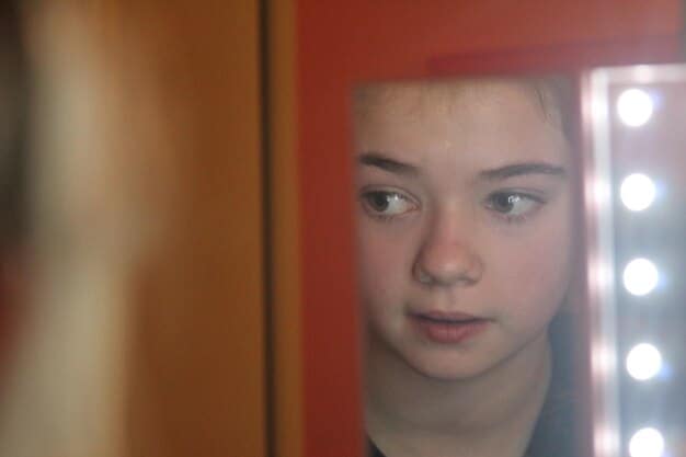 A close-up shot of Cassie Howard (Sydney Sweeney) crying in front of a mirror, surrounded by discarded clothing and makeup. The image captures her emotional breakdown and the pressures she faces to maintain her appearance and relationships.