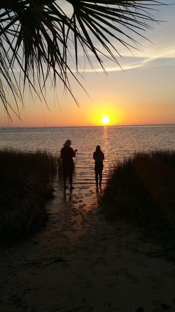A scenic image of Joey Graziadei and a contestant standing on a beach at sunset during the hometown visit in Hawaii. They are holding hands and looking out at the ocean, with the vibrant colors of the sunset reflecting in the water. The image conveys feelings of romance and serenity.