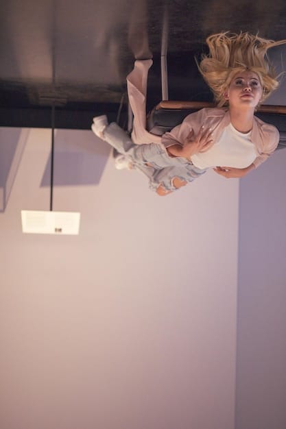 A medium shot of Asher, suspended upside down from the ceiling in his minimalist home. His face displays a mix of confusion, terror, and disbelief.