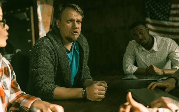 A medium shot of Kendall Roy (played by Jeremy Strong) in a high-powered business meeting. He is surrounded by other executives, and his expression is intense and determined. The background is a modern office setting with large windows overlooking a city skyline, emphasizing his ambition and drive.