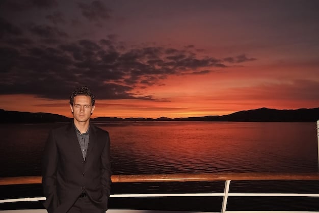 Roman Roy is standing alone on the deck of the yacht, looking out at the water with a mix of sadness and vulnerability on his face. The lighting is soft, highlighting the isolation of the character.
